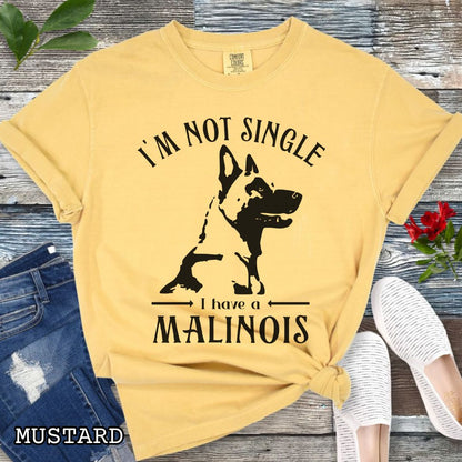 Mustard yellow Comfort Colors t-shirt with a woodcut-style Belgian Malinois graphic and the humorous text 'I'm Not Single, I Have a Malinois', shown on a wooden surface with accessories.