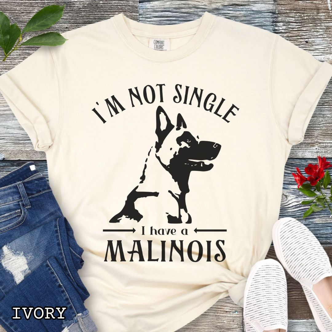 Beige Comfort Colors t-shirt with a woodcut-style Belgian Malinois graphic and the humorous text 'I'm Not Single, I Have a Malinois', shown on a wooden surface with accessories.