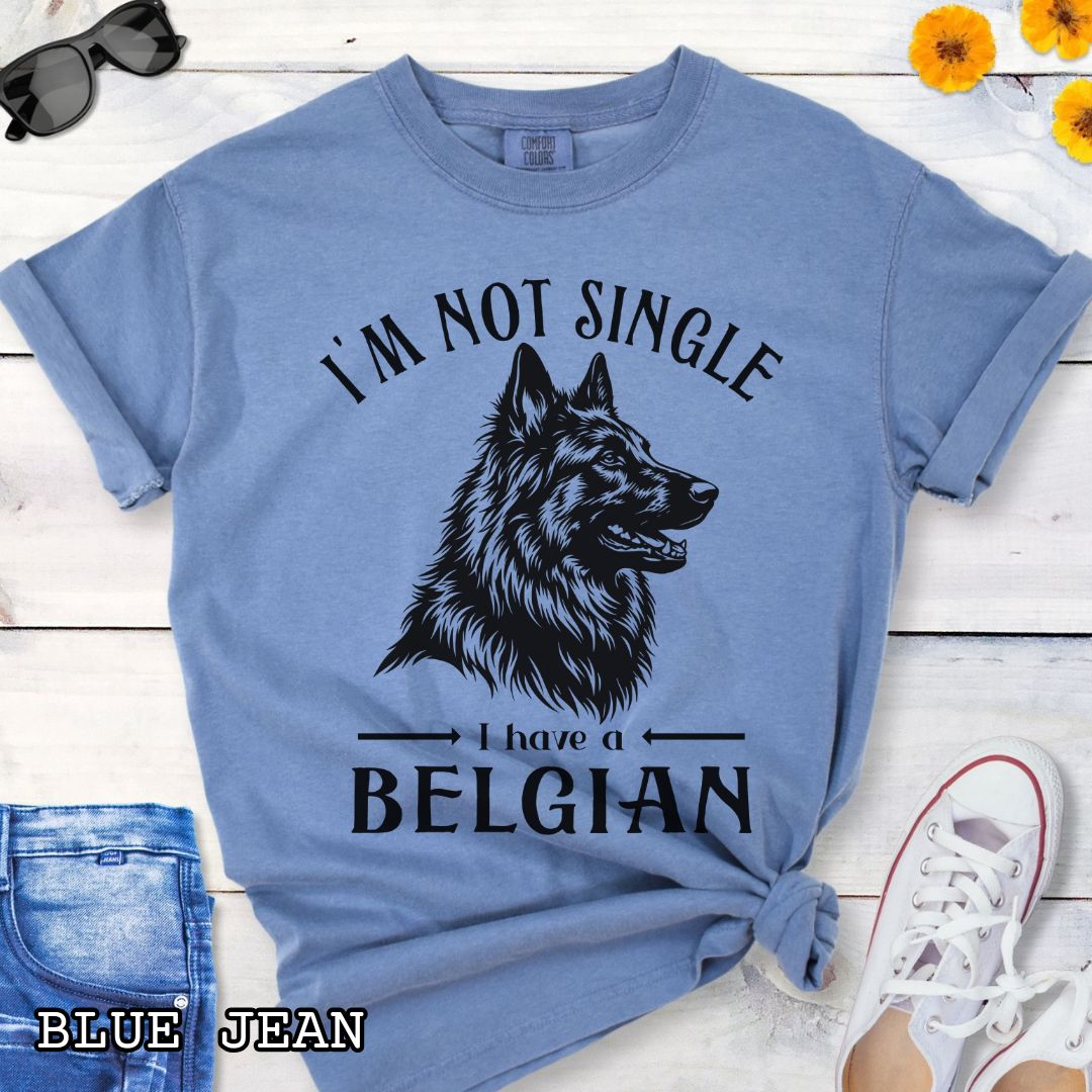 Blue Comfort Colors t-shirt with a woodcut-style Belgian Sheepdog graphic and humorous text 'I'm Not Single, I Have a Belgian', shown on a wooden surface with jeans and sneakers.