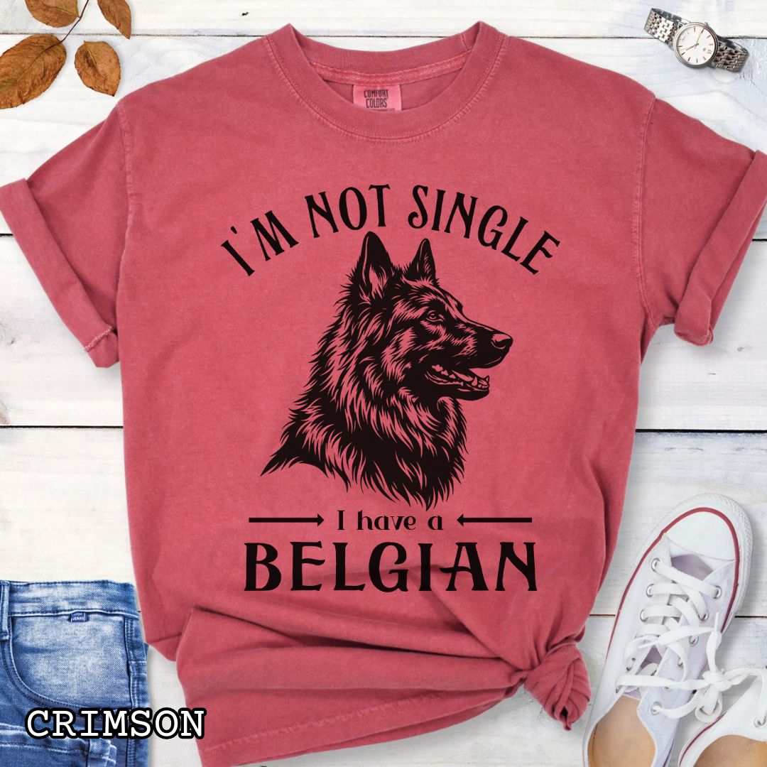 Red Comfort Colors t-shirt with a woodcut-style Belgian Sheepdog graphic and humorous text 'I'm Not Single, I Have a Belgian', shown on a wooden surface with jeans and sneakers.