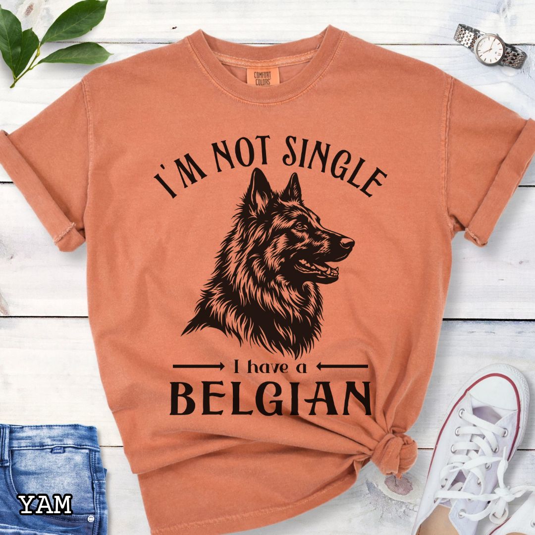 Orange Comfort Colors t-shirt with a woodcut-style Belgian Sheepdog graphic and humorous text 'I'm Not Single, I Have a Belgian', shown on a wooden surface with jeans and sneakers.