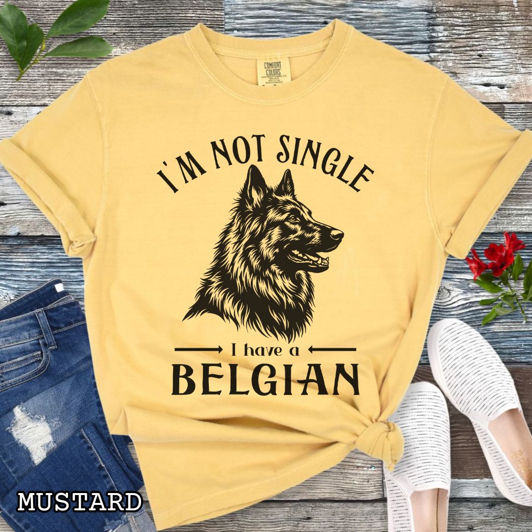 Mustard yellow Comfort Colors t-shirt with a woodcut-style Belgian Sheepdog graphic and humorous text 'I'm Not Single, I Have a Belgian', shown on a wooden surface with jeans and sneakers.