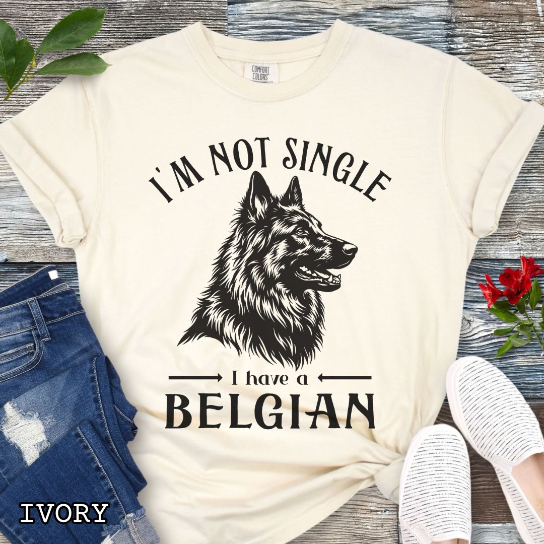 Beige Comfort Colors t-shirt with a woodcut-style Belgian Sheepdog graphic and humorous text 'I'm Not Single, I Have a Belgian', shown on a wooden surface with jeans and sneakers.