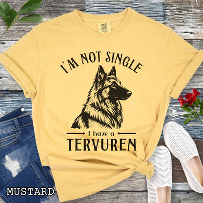 Mustard yellow Comfort Colors t-shirt with a woodcut-style Belgian Tervuren graphic and humorous text 'I'm Not Single, I Have a Belgian', shown on a wooden surface with jeans and sneakers.