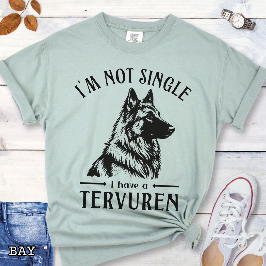 Light green Comfort Colors t-shirt with a woodcut-style Belgian Tervuren graphic and humorous text 'I'm Not Single, I Have a Belgian', shown on a wooden surface with jeans and sneakers.