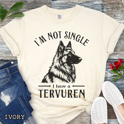Beige Comfort Colors t-shirt with a woodcut-style Belgian Tervuren graphic and humorous text 'I'm Not Single, I Have a Belgian', shown on a wooden surface with jeans and sneakers.