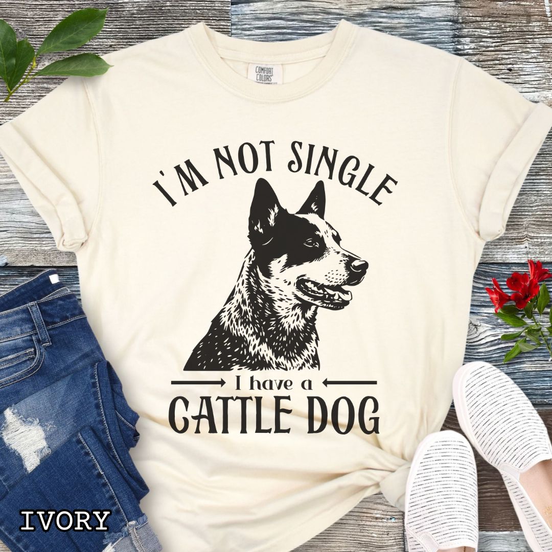 Beige Comfort Colors t-shirt with a woodcut-style graphic of an Australian Cattle Dog and humorous text 'I'm not single, I have a cattle dog', shown on a wooden surface with jeans and shoes.