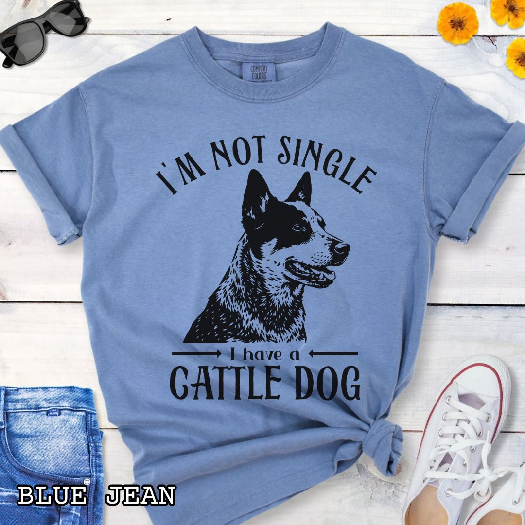 Blue Comfort Colors t-shirt with a woodcut-style graphic of an Australian Cattle Dog and humorous text 'I'm not single, I have a cattle dog', shown on a wooden surface with jeans and shoes.
