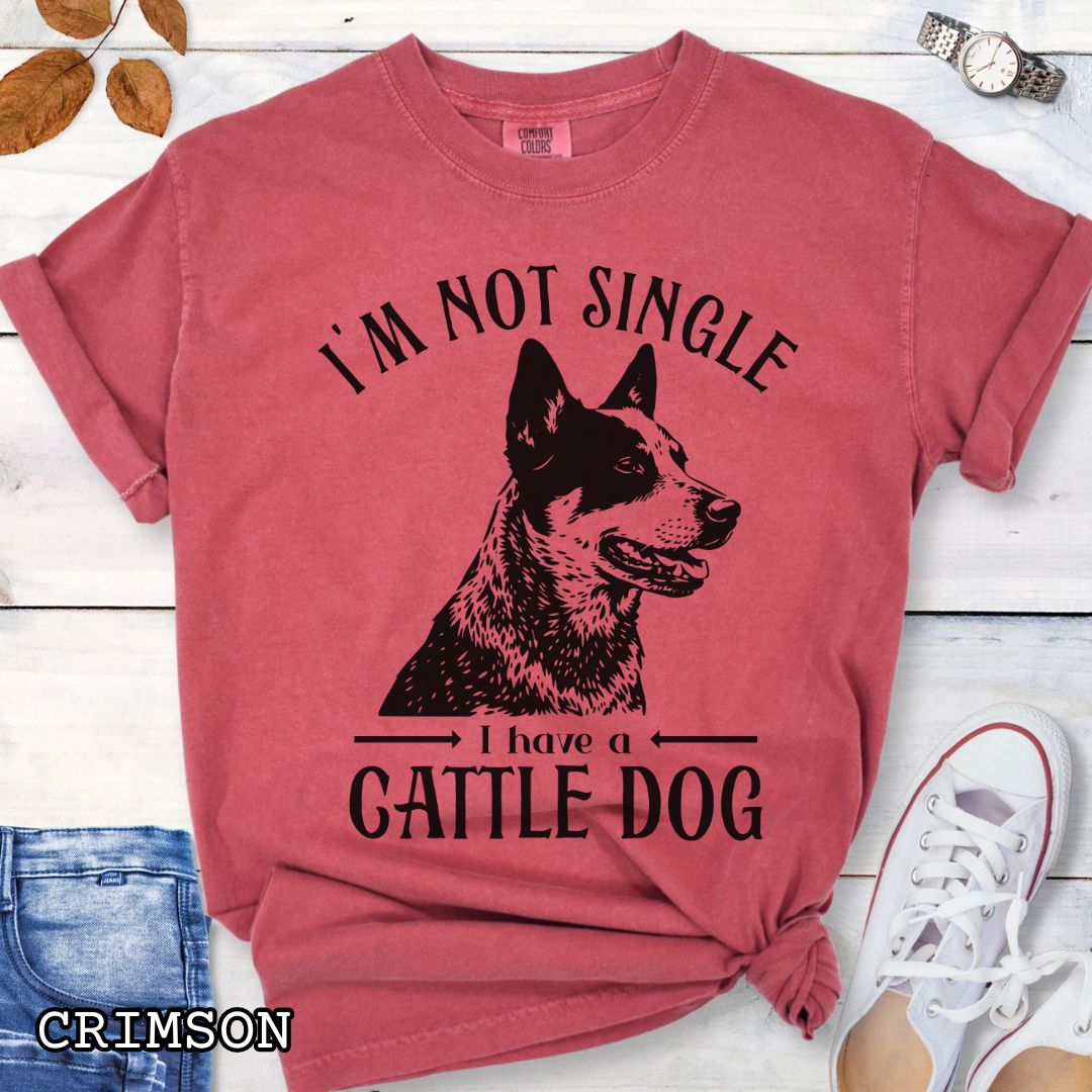 Red Comfort Colors t-shirt with a woodcut-style graphic of an Australian Cattle Dog and humorous text 'I'm not single, I have a cattle dog', shown on a wooden surface with jeans and shoes.