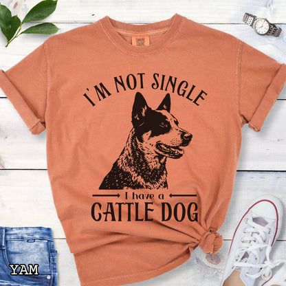 Orange Comfort Colors t-shirt with a woodcut-style graphic of an Australian Cattle Dog and humorous text 'I'm not single, I have a cattle dog', shown on a wooden surface with jeans and shoes.