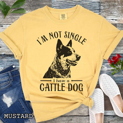 Mustard yellow Comfort Colors t-shirt with a woodcut-style graphic of an Australian Cattle Dog and humorous text 'I'm not single, I have a cattle dog', shown on a wooden surface with jeans and shoes.