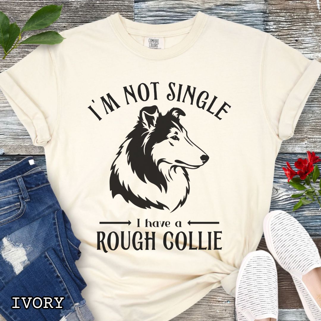 Beige Comfort Colors t-shirt with a Rough Collie graphic and humorous text 'I'm Not Single, I Have a Rough Collie', shown on a wooden surface with accessories.