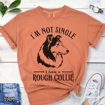 Orange Comfort Colors t-shirt with a Rough Collie graphic and humorous text 'I'm Not Single, I Have a Rough Collie', shown on a wooden surface with accessories.