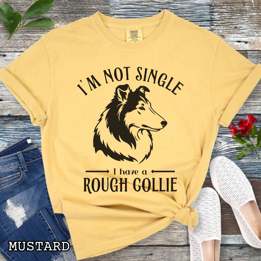 Mustard yellow Comfort Colors t-shirt with a Rough Collie graphic and humorous text 'I'm Not Single, I Have a Rough Collie', shown on a wooden surface with accessories.