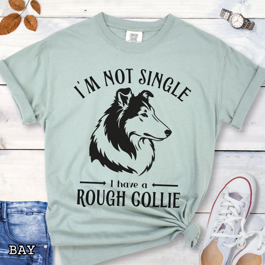 Light green Comfort Colors t-shirt with a Rough Collie graphic and humorous text 'I'm Not Single, I Have a Rough Collie', shown on a wooden surface with accessories.