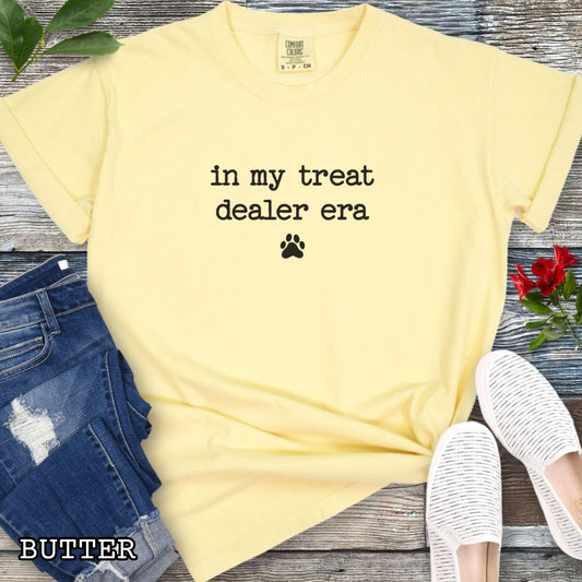 Yellow Comfort Colors t-shirt for dog lovers with humorous text 'in my treat dealer era' in typewriter font, and a small paw print, shown on a wooden surface with jeans and shoes.