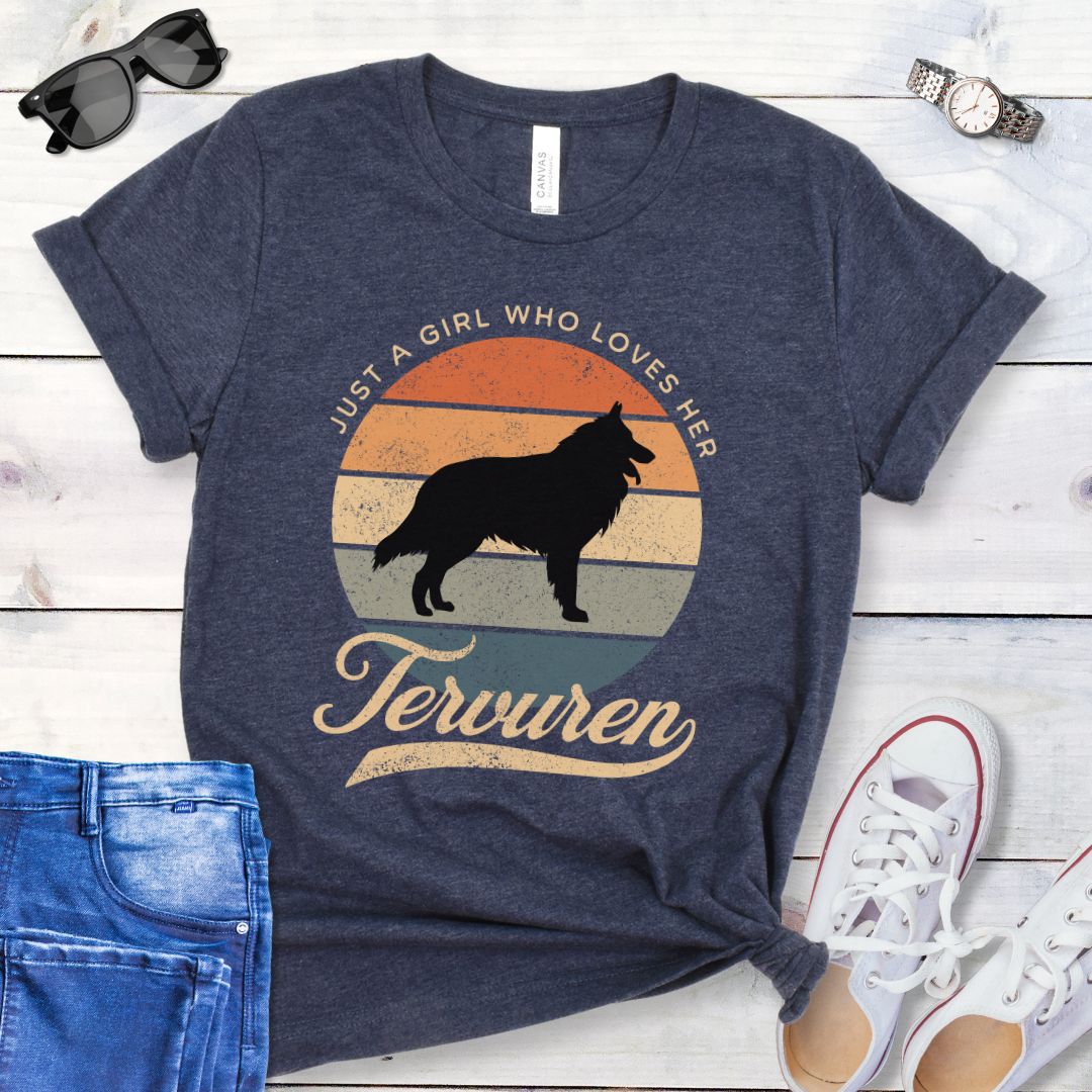 Heather navy blue t-shirt for Belgian Tervuren lovers with vintage sunset graphic and Tervuren dog silhouette, and text 'Just a Girl Who Loves Her Tervuren', shown on a wooden surface with accessories.
