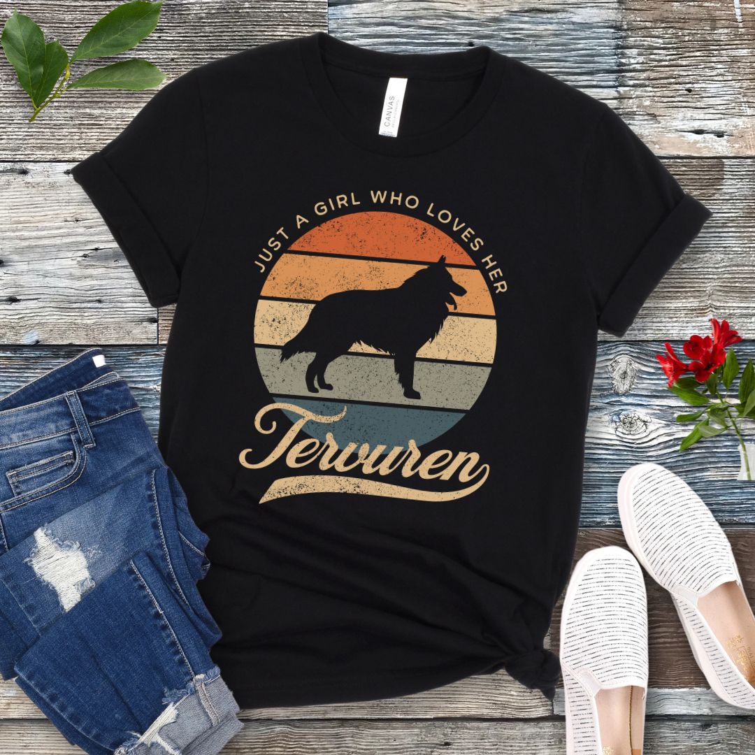 Black t-shirt for Belgian Tervuren lovers with vintage sunset graphic and Tervuren dog silhouette, and text 'Just a Girl Who Loves Her Tervuren', shown on a wooden surface with accessories.