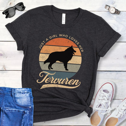 Dark grey t-shirt for Belgian Tervuren lovers with vintage sunset graphic and Tervuren dog silhouette, and text 'Just a Girl Who Loves Her Tervuren', shown on a wooden surface with accessories.