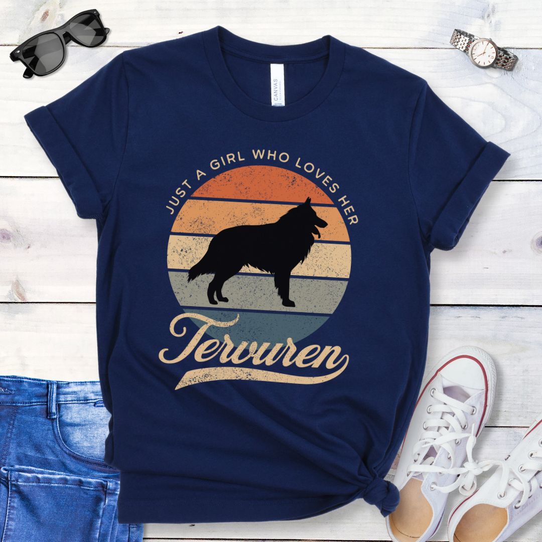 Navy blue t-shirt for Belgian Tervuren lovers with vintage sunset graphic and Tervuren dog silhouette, and text 'Just a Girl Who Loves Her Tervuren', shown on a wooden surface with accessories.