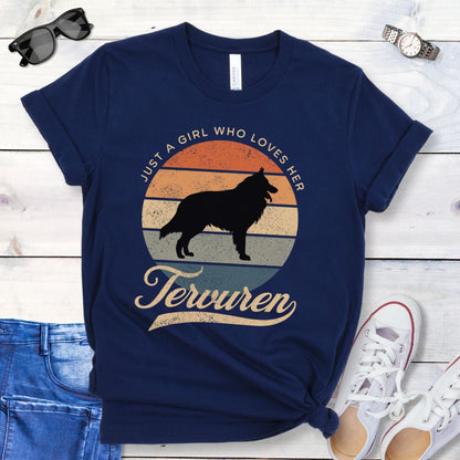 Navy blue t-shirt for Belgian Tervuren lovers with vintage sunset graphic and Tervuren dog silhouette, and text 'Just a Girl Who Loves Her Tervuren', shown on a wooden surface with accessories.