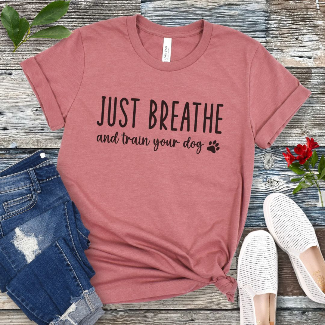 Mauve unisex dog training t-shirt with 'Just Breathe and train your dog' text and paw print, presented on a wooden surface with jeans and shoes.