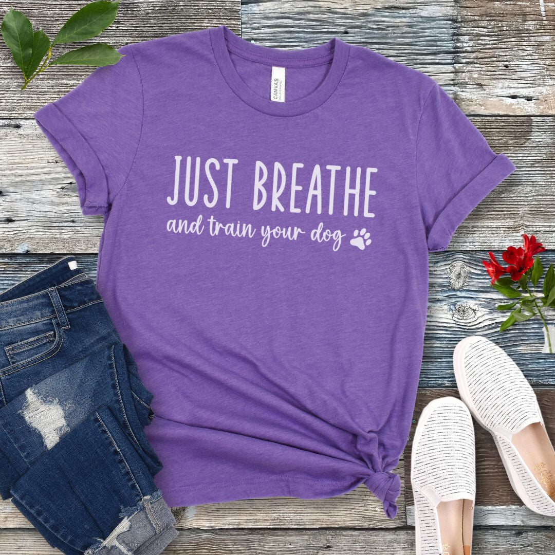 Purple unisex dog training t-shirt with 'Just Breathe and train your dog' text and paw print, presented on a wooden surface with jeans and shoes.