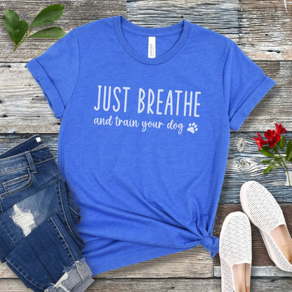 Royal blue unisex dog training t-shirt with 'Just Breathe and train your dog' text and paw print, presented on a wooden surface with jeans and shoes.