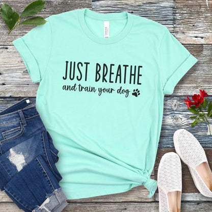 Mint green unisex dog training t-shirt with 'Just Breathe and train your dog' text and paw print, presented on a wooden surface with jeans and shoes.