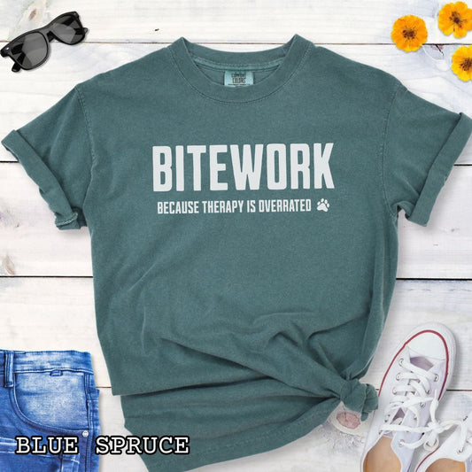 Green Comfort Colors t-shirt for k9 bite sports enthusiasts with humorous text 'Bitework because therapy is overrated', shown on a wooden surface with sunglasses and jeans.