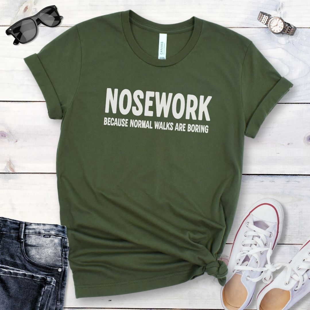 Military green K9 Nose Work t-shirt with 'NOSEWORK BECAUSE NORMAL WALKS ARE BORING' text on a wooden surface with jeans and shoes.