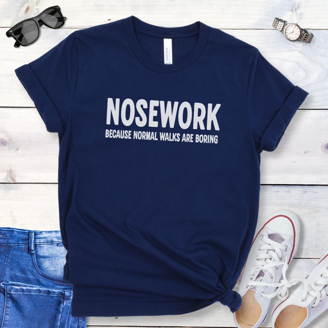 Navy blue K9 Nose Work t-shirt with 'NOSEWORK BECAUSE NORMAL WALKS ARE BORING' text on a wooden surface with jeans and shoes.