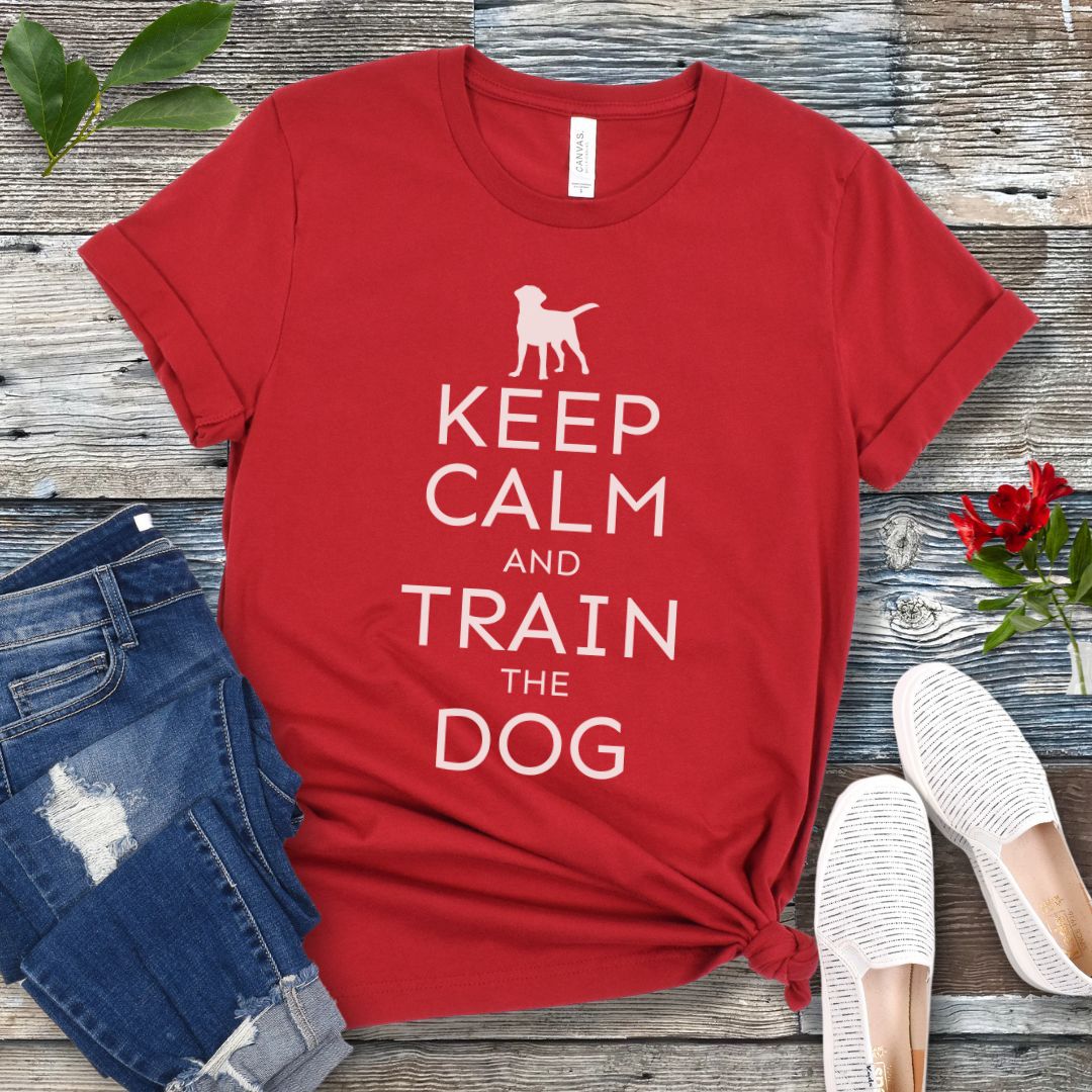 A red t-shirt with white text and dog graphic and text that says 'KEEP CALM AND TRAIN THE DOG', displayed on a wooden background with a pair of blue jeans and white shoes.
