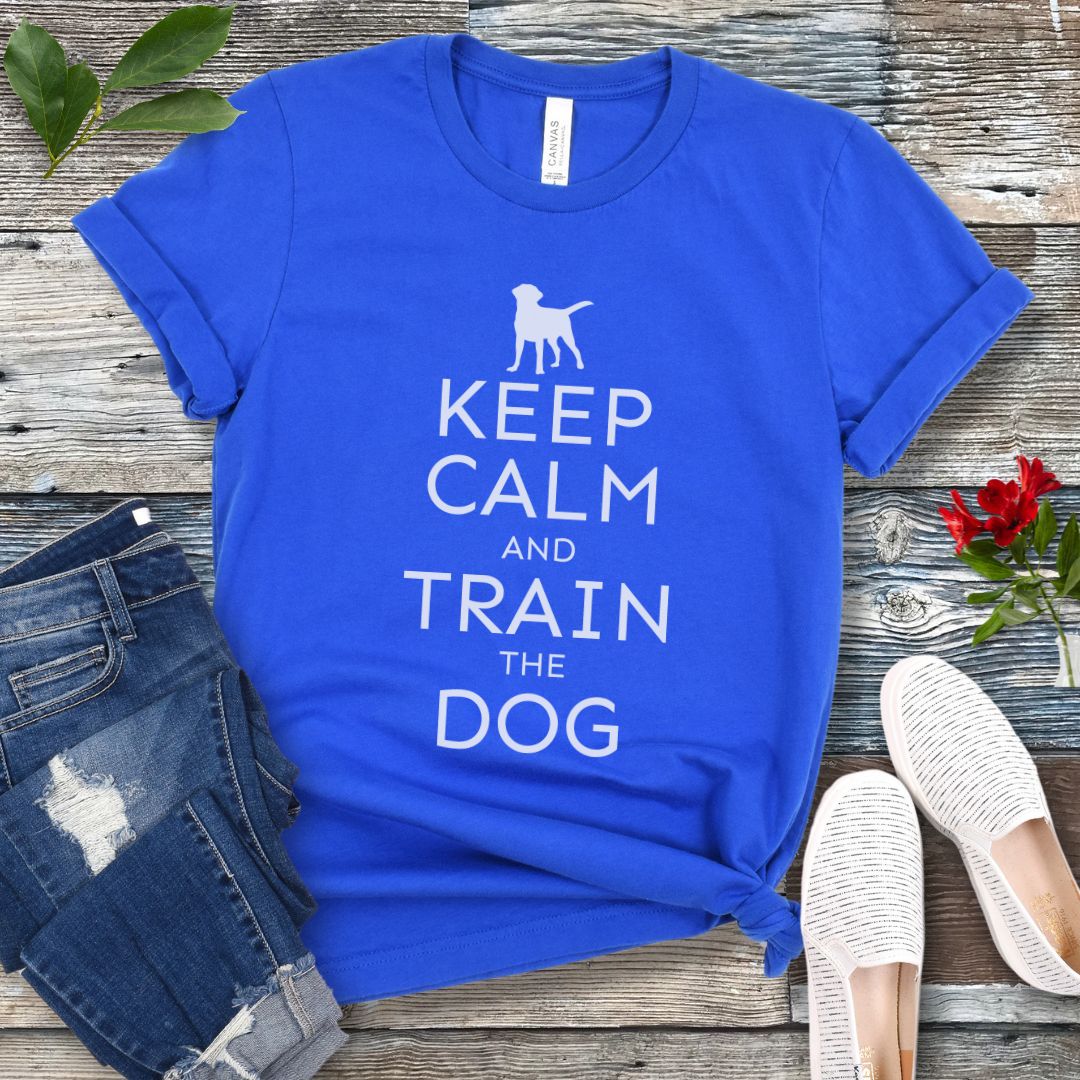 A royal blue t-shirt with white text and dog graphic and text that says 'KEEP CALM AND TRAIN THE DOG', displayed on a wooden background with a pair of blue jeans and white shoes.