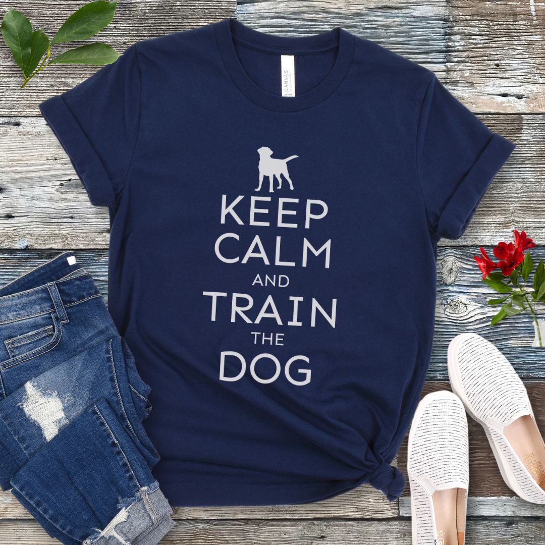 A navy blue t-shirt with white text and dog graphic and text that says 'KEEP CALM AND TRAIN THE DOG', displayed on a wooden background with a pair of blue jeans and white shoes.