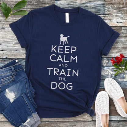 A navy blue t-shirt with white text and dog graphic and text that says 'KEEP CALM AND TRAIN THE DOG', displayed on a wooden background with a pair of blue jeans and white shoes.