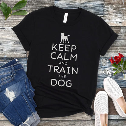 A black t-shirt with white text and dog graphic and text that says 'KEEP CALM AND TRAIN THE DOG', displayed on a wooden background with a pair of blue jeans and white shoes.