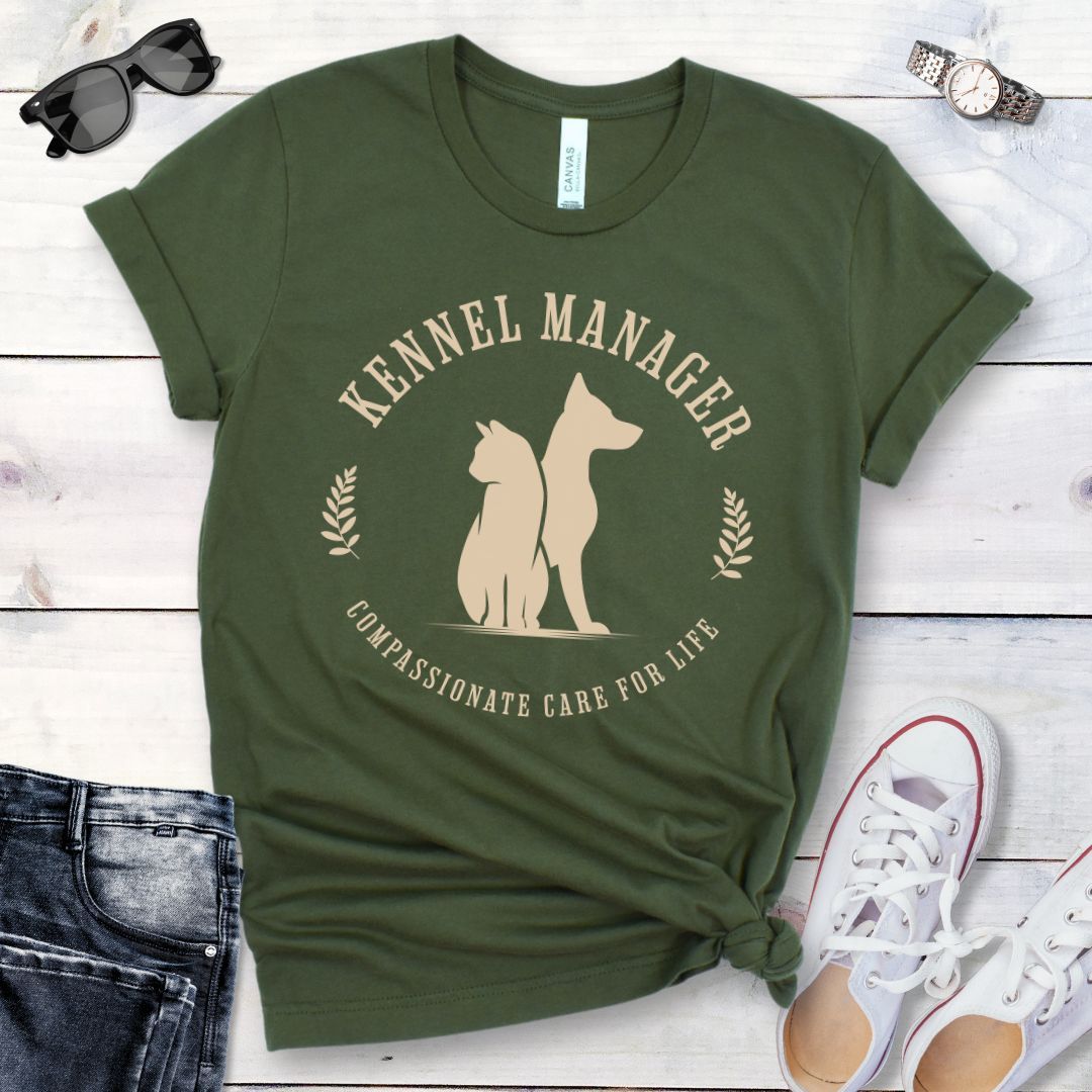 A military green t-shirt with 'Kennel Manager' text and a graphic design of a dog and cat, placed flat with a pair of jeans and white shoes on a wooden surface.