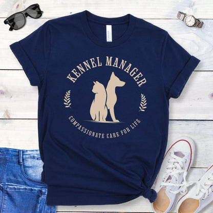 A navy blue t-shirt with 'Kennel Manager' text and a graphic design of a dog and cat, placed flat with a pair of jeans and white shoes on a wooden surface.