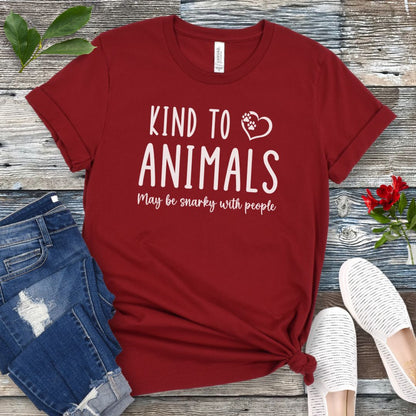 Red unisex t-shirt for pet and animal lovers with humorous text 'Kind to Animals May be snarky with people' in playful lettering, shown on a wooden surface with accessories.