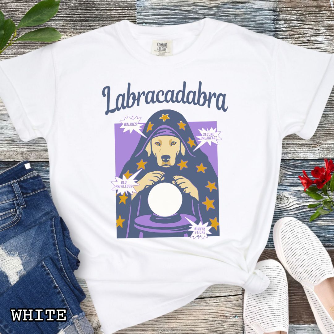 A white short-sleeve graphic T-shirt laid flat on a rustic wood surface. The shirt graphic reads 'Labracadabra' above an illustration of a Labrador Retriever dog dressed as a witch holding a crystal ball, with purple and yellow tones.