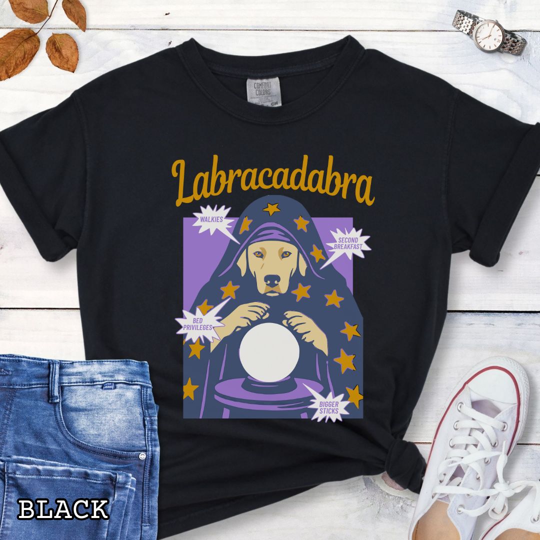 A black short-sleeve graphic T-shirt laid flat on a rustic wood surface. The shirt graphic reads 'Labracadabra' above an illustration of a Labrador Retriever dog dressed as a witch holding a crystal ball, with purple and yellow tones.