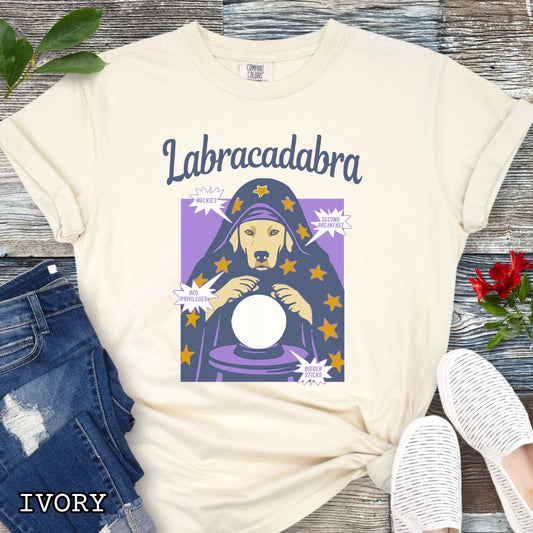 An ivory short-sleeve graphic T-shirt laid flat on a rustic wood surface. The shirt graphic reads 'Labracadabra' above an illustration of a Labrador Retriever dog dressed as a witch holding a crystal ball, with purple and yellow tones.