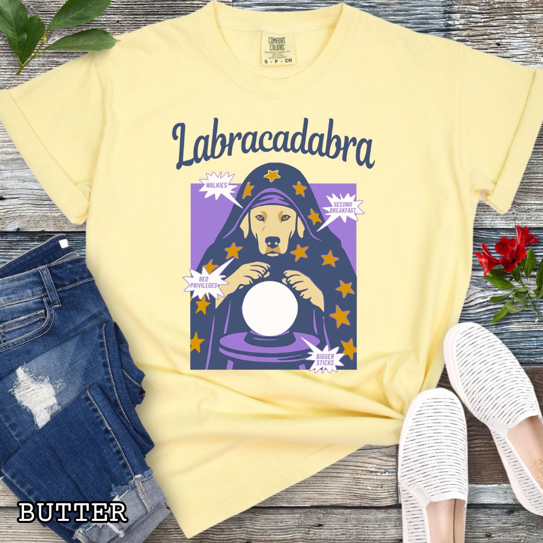 A yellow short-sleeve graphic T-shirt laid flat on a rustic wood surface. The shirt graphic reads 'Labracadabra' above an illustration of a Labrador Retriever dog dressed as a witch holding a crystal ball, with purple and yellow tones.