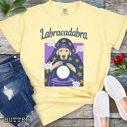 A yellow short-sleeve graphic T-shirt laid flat on a rustic wood surface. The shirt graphic reads 'Labracadabra' above an illustration of a Labrador Retriever dog dressed as a witch holding a crystal ball, with purple and yellow tones.