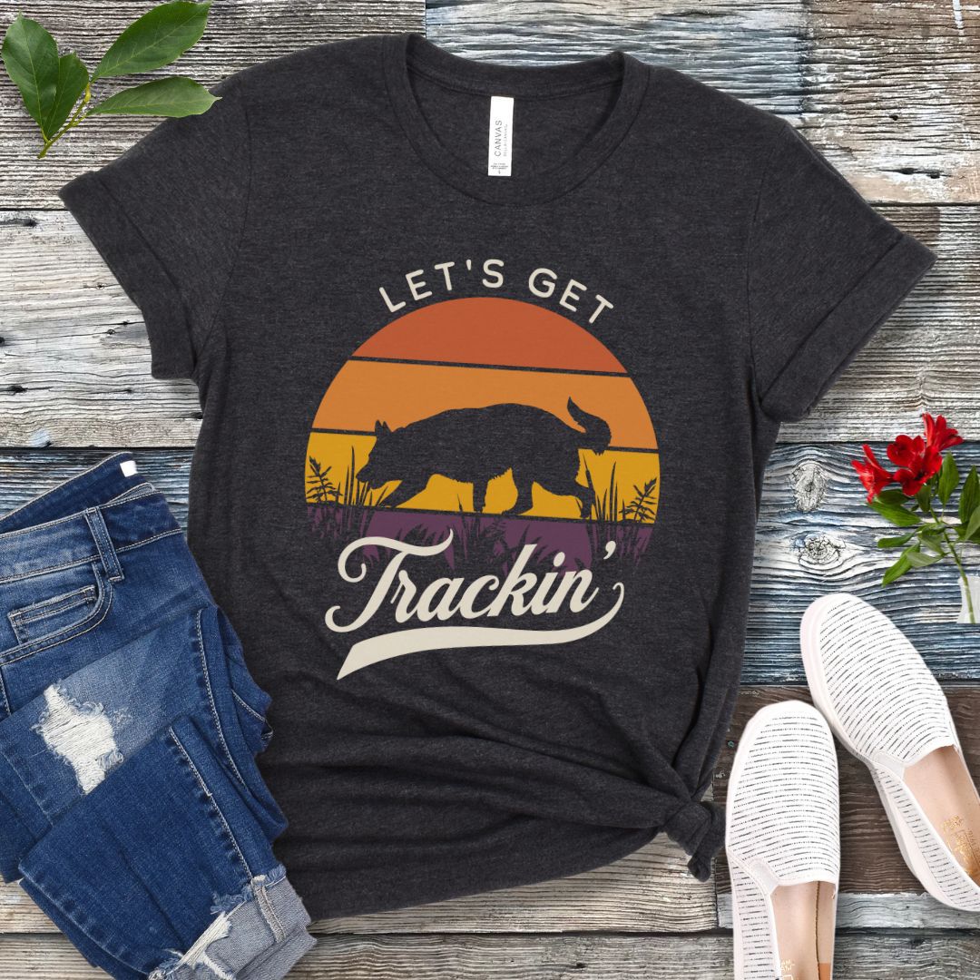 A dark grey t-shirt with the text 'LET'S GET Trackin'' with a custom breed tracking dog silhouette against a vintage sunset backdrop, displayed on a wooden surface with jeans and white shoes in the surrounding area.