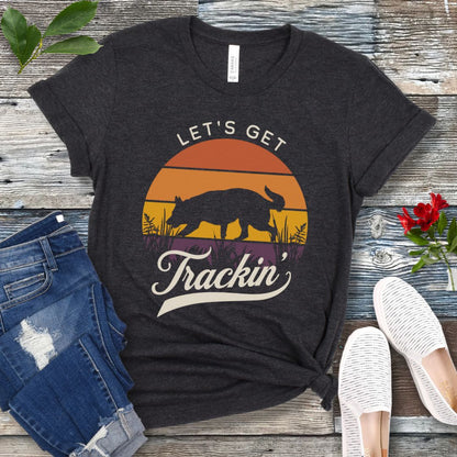 A dark grey t-shirt with the text 'LET'S GET Trackin'' with a custom breed tracking dog silhouette against a vintage sunset backdrop, displayed on a wooden surface with jeans and white shoes in the surrounding area.