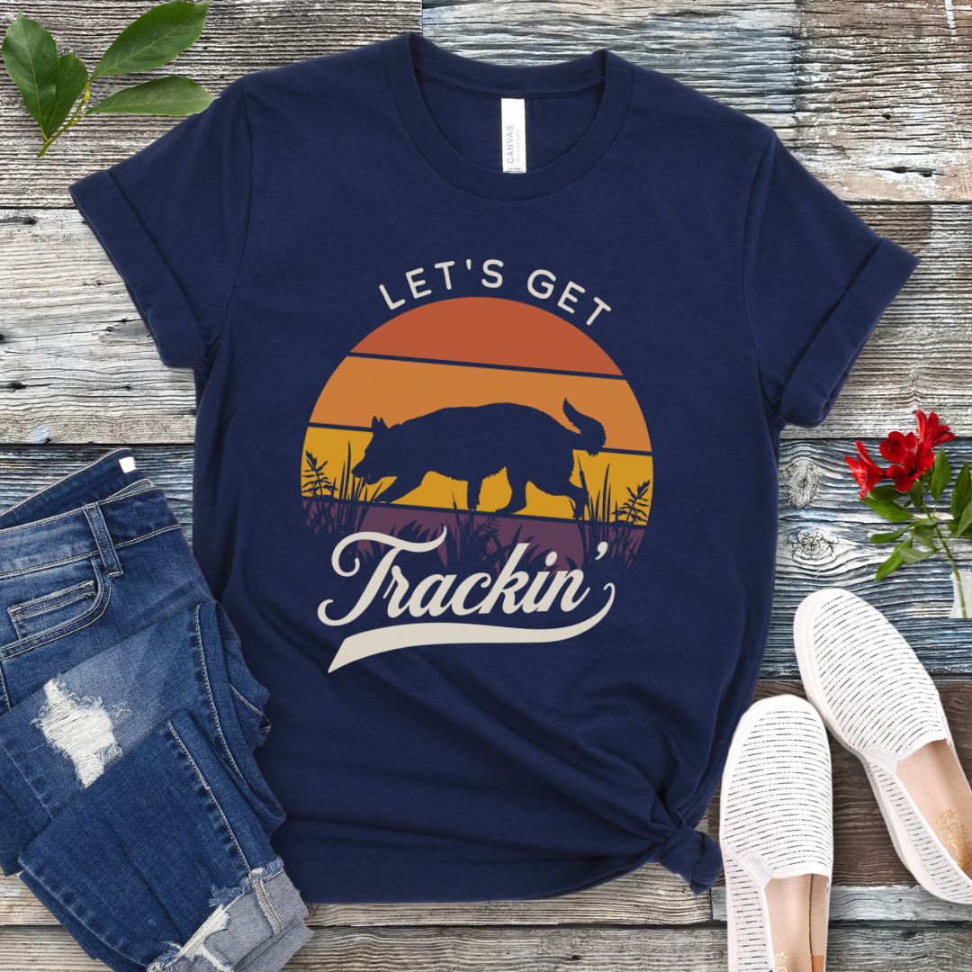 A navy t-shirt with the text 'LET'S GET Trackin'' with a custom breed tracking dog silhouette against a vintage sunset backdrop, displayed on a wooden surface with jeans and white shoes in the surrounding area.