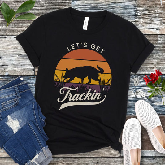 A black t-shirt with the text 'LET'S GET Trackin'' and a custom tracking dog silhouette against a vintage sunset backdrop, displayed on a wooden surface with jeans and white shoes in the surrounding area.
