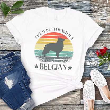 White t-shirt for Belgian Sheepdog lovers with a retro sunset graphic design featuring a Belgian Sheepdog silhouette and text 'Life is better with a Belgian', shown on a wooden surface.